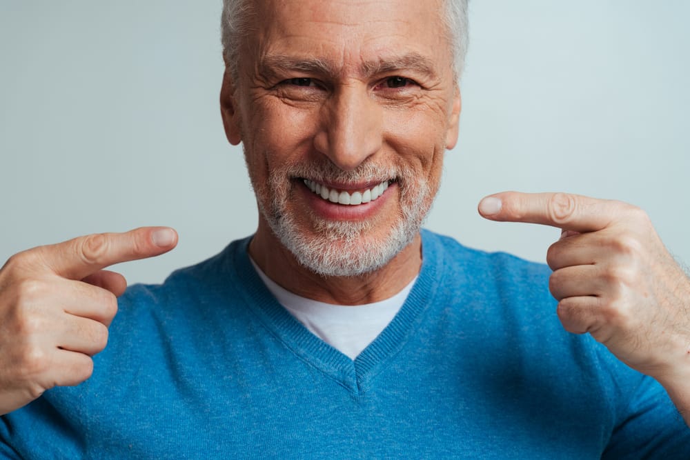 Senior Man Pointing to Healthy Smile - Fillings Happy senior man proudly pointing to his bright, healthy smile, showcasing excellent dental care results. - Fillings