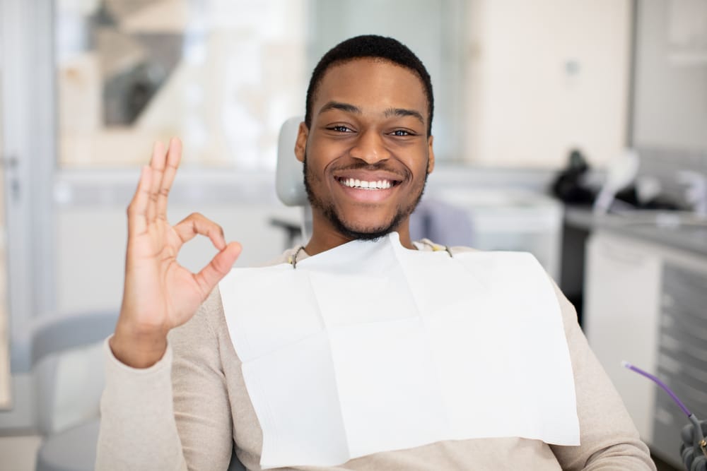 Smiling man giving OK sign at the dentist - Cosmetic Dentistry A cheerful man sits in the dental chair, wearing a protective bib and giving an OK hand gesture after his cosmetic dental treatment - Cosmetic Dentistry.