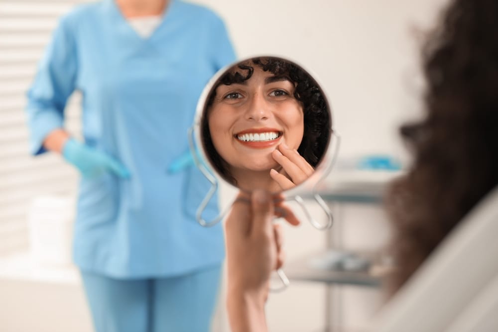Smiling woman looking at her teeth in a dental mirror after a successful treatment, showcasing a healthy smile. - Veneers