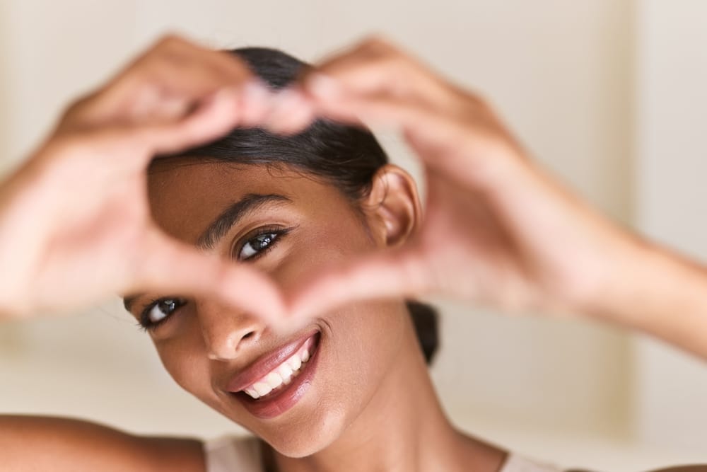 Woman making a heart shape with her hands and smiling - Cosmetic Dentistry A young woman forms a heart shape with her hands and beams a perfect smile, showcasing the results of professional cosmetic dental care - Cosmetic Dentistry.