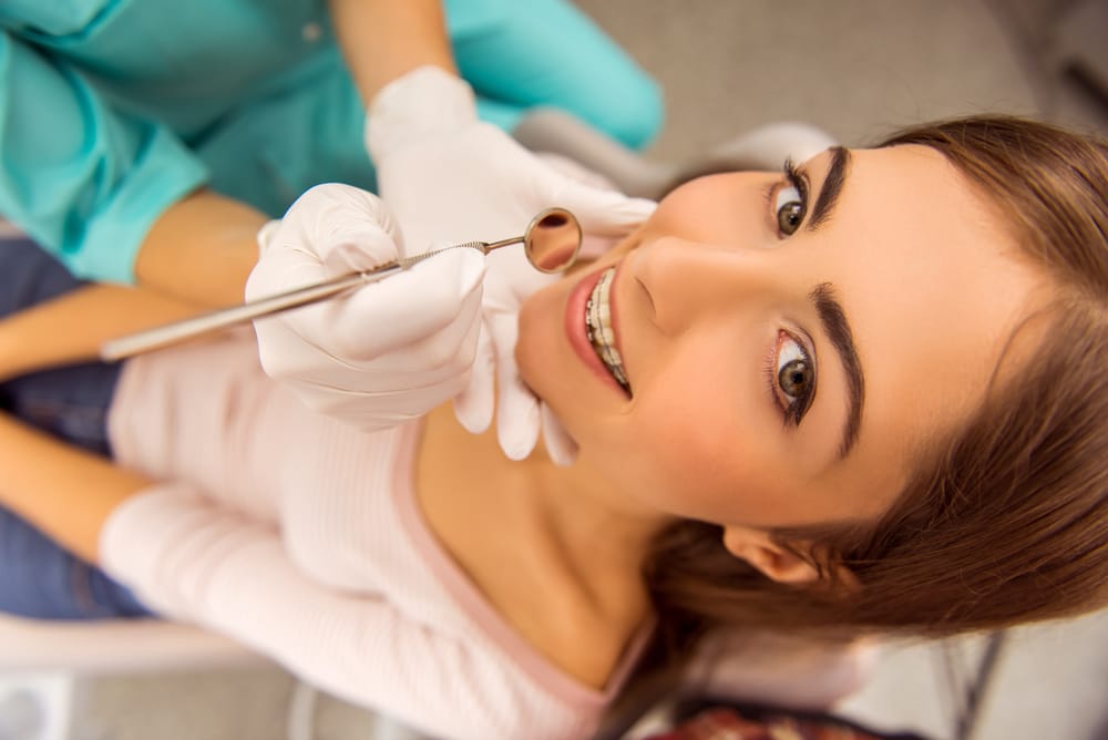 A young girl with braces looks up and smiles during a dental check-up as a dentist holds a mirror tool to examine her teeth - Braces.