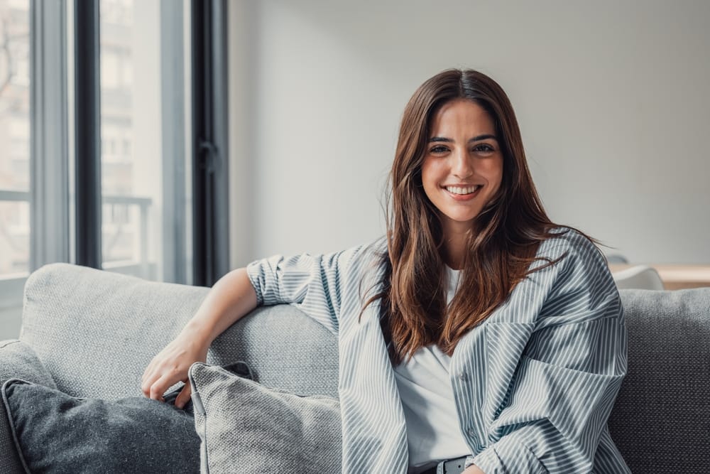 Confident woman with a bright smile at home - Cosmetic Dentistry A smiling woman with long brown hair sits comfortably on a couch, showing off her natural, radiant smile that reflects confidence and happiness - Cosmetic Dentistry.