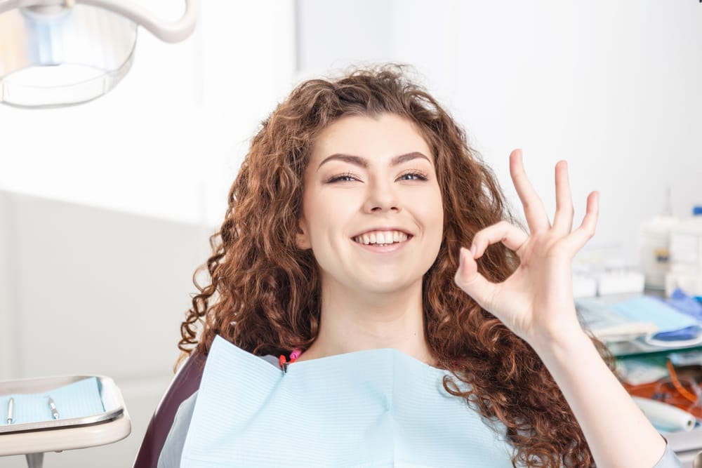 Smiling woman in a dental chair giving an OK hand gesture after a positive and comfortable dental visit - Periodontal Care