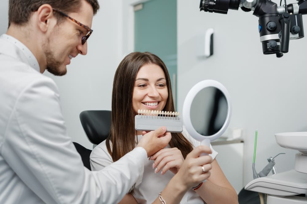 Smiling woman comparing tooth shades with her dentist to choose the perfect color for dental restoration or whitening. - Veneers