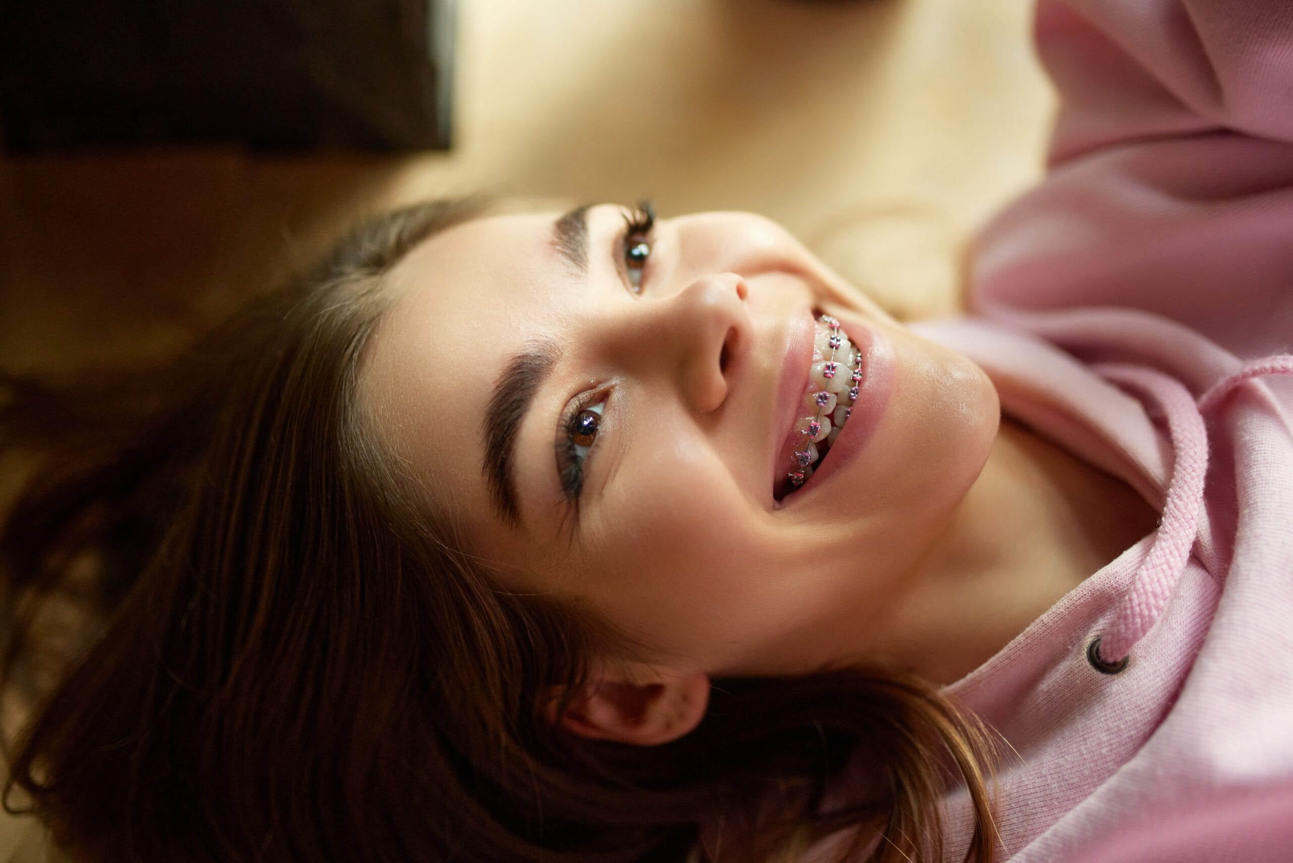 A close-up of a joyful young girl lying down in a pink hoodie, showing off her colorful braces with a bright, carefree smile - Braces.