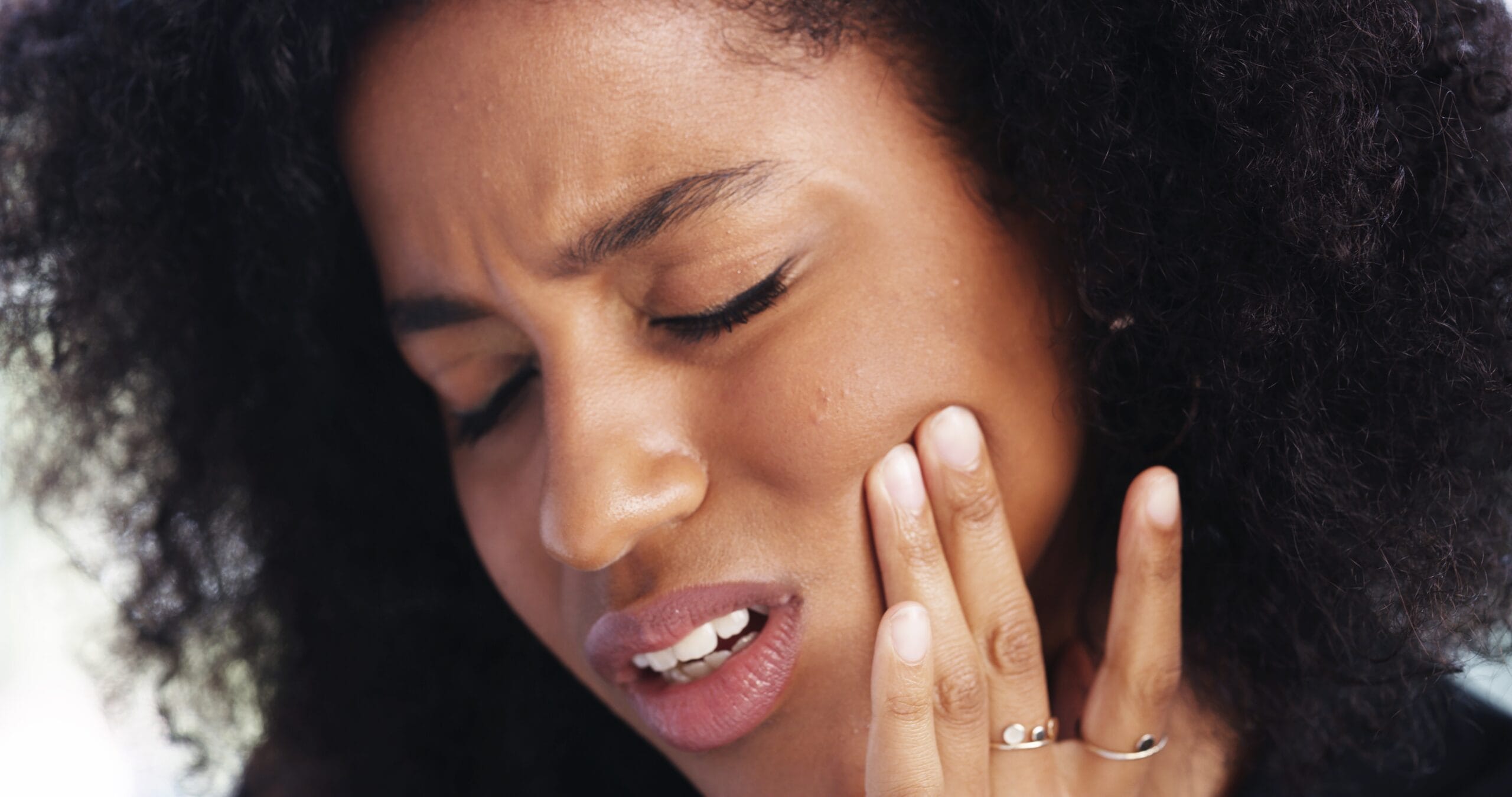 Young woman touching her cheek with a pained expression, showing symptoms of toothache or dental sensitivity. - TMJ