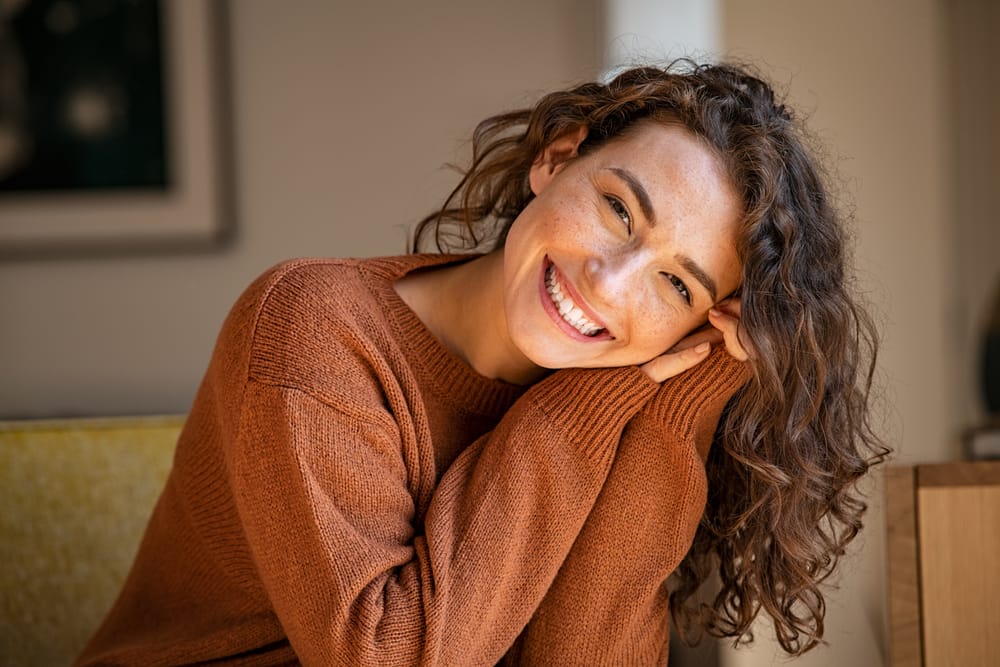 Cheerful woman in a warm sweater smiling with joy, reflecting confidence and excellent oral health. - Teeth Whitening